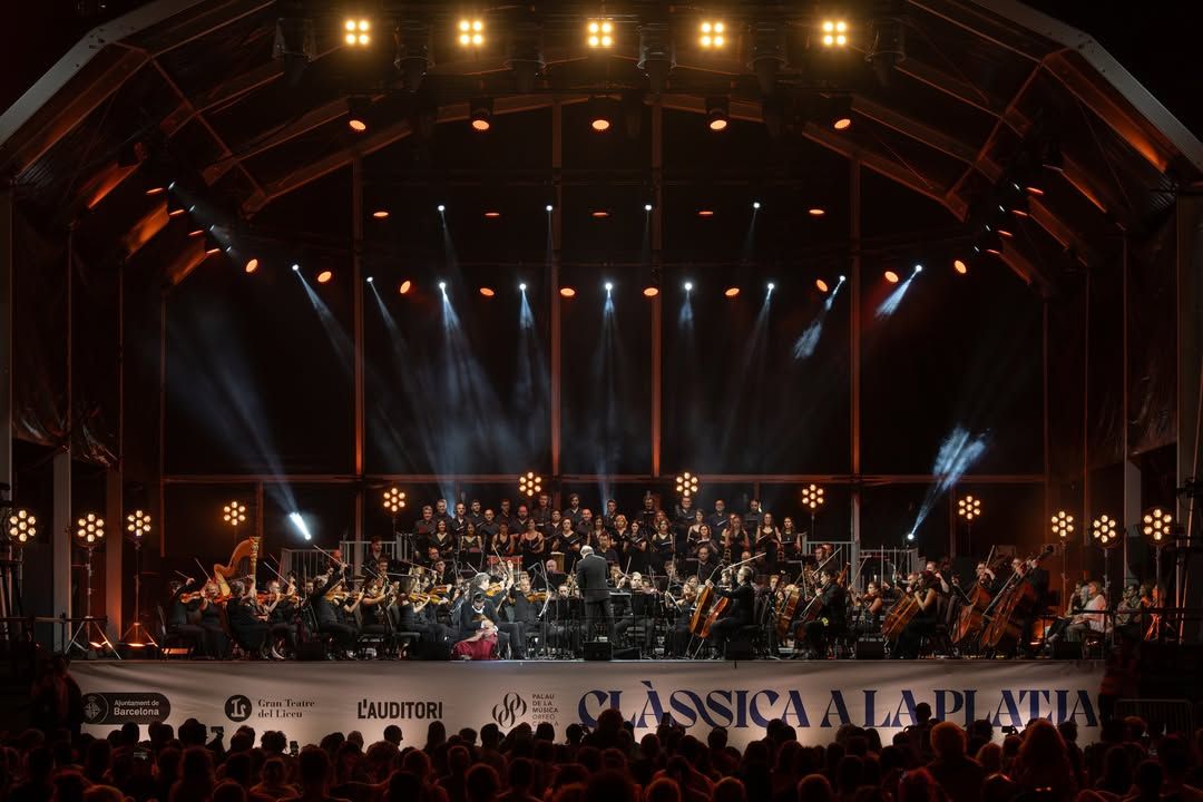Foto de la orquesta interpretando sobre el escenario Orbit 25 de Homs Rentals, con el coro situado en los niveles elevados de tarima y el público en la playa disfrutando de la música sinfónica.