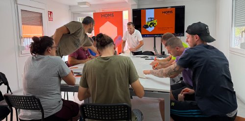 Group of adults at a workshop around a table, listening to a presenter at the front with two screens showing a slide.